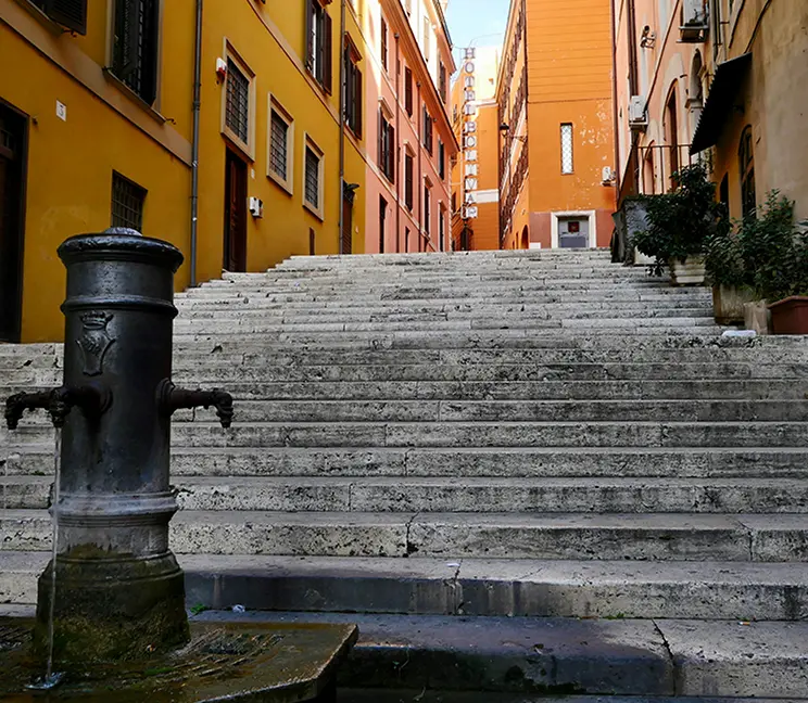 Fuente pública en un callejón romano, con escaleras que conducen a edificios coloridos al fondo.