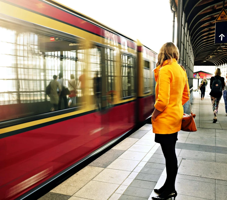 Donna in cappotto arancione sul marciapiede di una stazione, mentre un treno rosso sfreccia davanti a lei